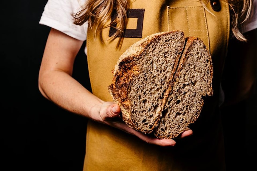 A woman holds a loaf of bread, she is wearing a mustard-coloured apron.