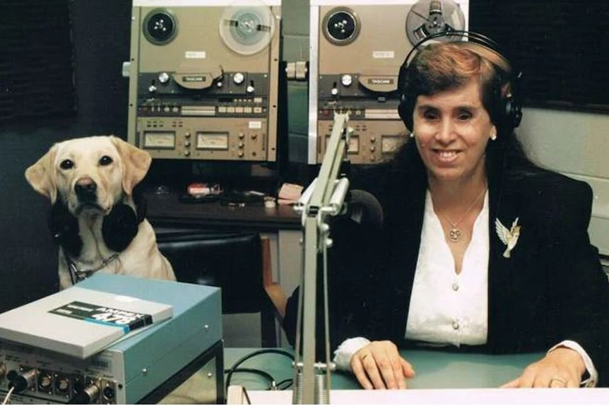 Elaine sits behind a radio broadcasting microphone. She is smiling and wearing headphone. Her guide dog also wears headphones.