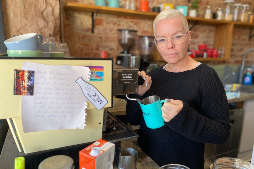 A woman with white hair makes a coffee at a machine while holding a green jug