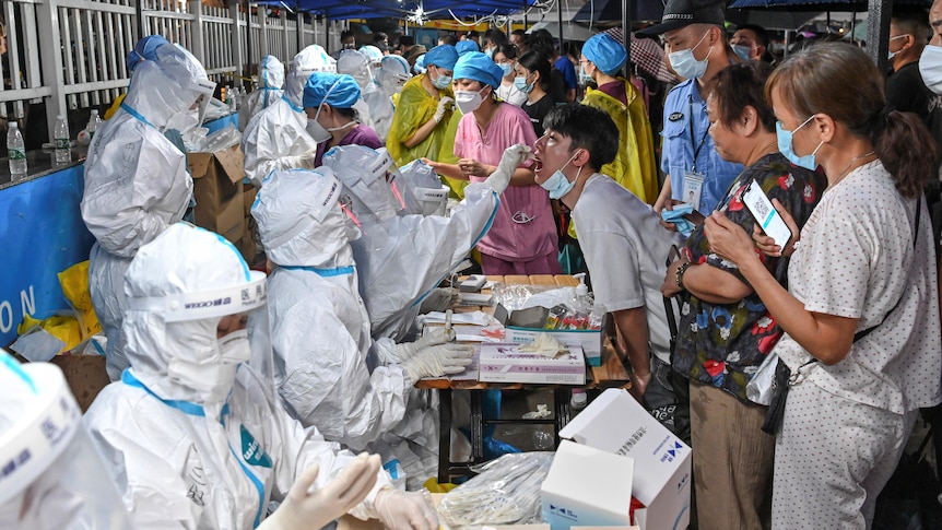 A young man lowers a face mask so one of many health workers in full PPE can swab his throat at a bust testing centre.