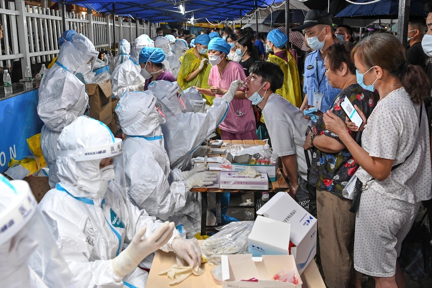 A young man lowers a face mask so one of many health workers in full PPE can swab his throat at a bust testing centre.