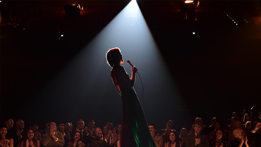 A woman standing on stage holding a microphone with a spotlight on her and the audience behind