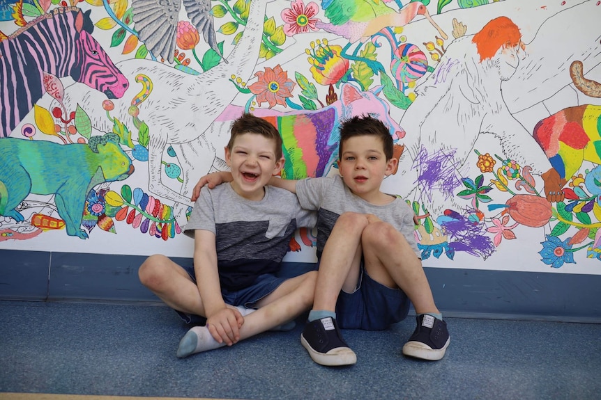 One boy smiling, one serious arms around each other sitting on floor in front of colourful mural