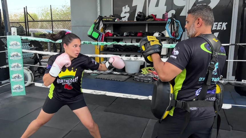 A girl in pink boxing gloves prepares to swing at a man in protective gear.