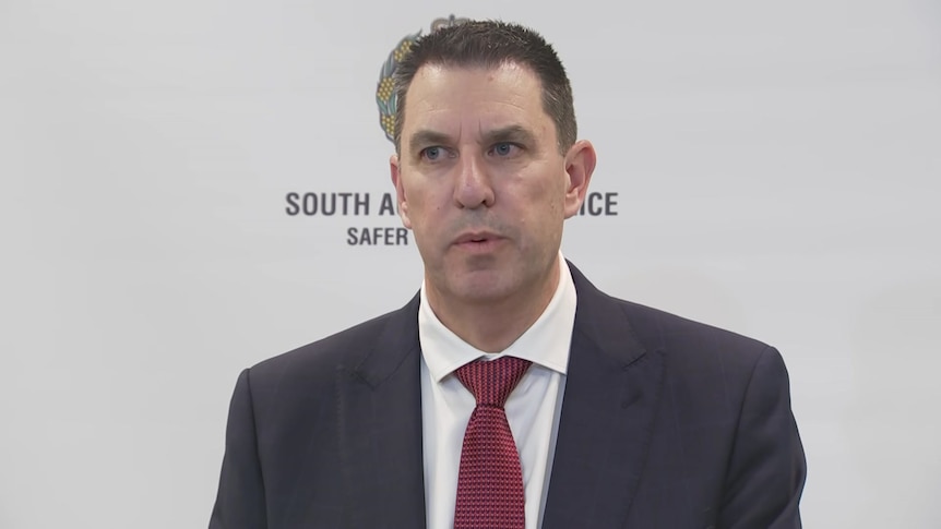 a man in a business suit and red tie speaking in front of sa police banner