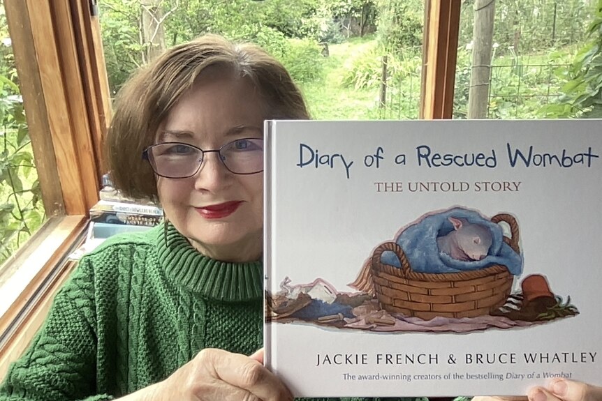 Jackie French wearing glasses and a green sweater holding up a copy of one of her books