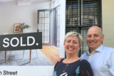 A middle aged couple stand in front of a sold sign