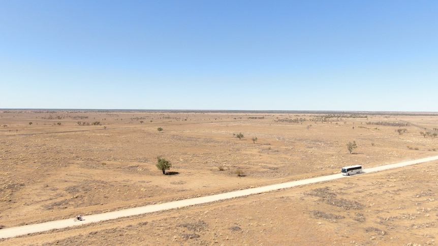 A man on a motorbike rides in front of a school bus surrounded by a parched landscape under a blue cloudless sky