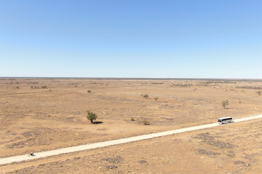 A man on a motorbike rides in front of a school bus surrounded by a parched landscape under a blue cloudless sky