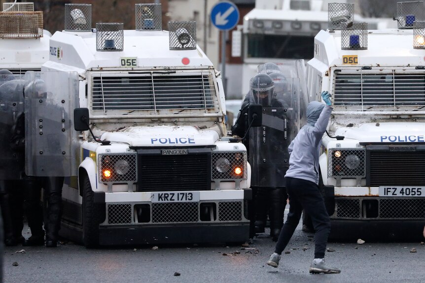 A hooded person goes to throw something at a riot police officer