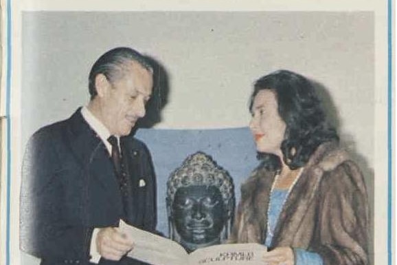 A man in a suit and a woman in fur coat hold an exhibition catalogue in front of a sculpture