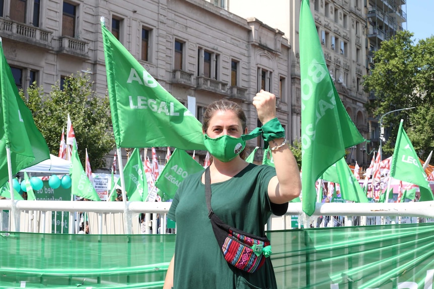A woman wearing a green mask and shirt and a tie around her wrist stands in front of green flags.