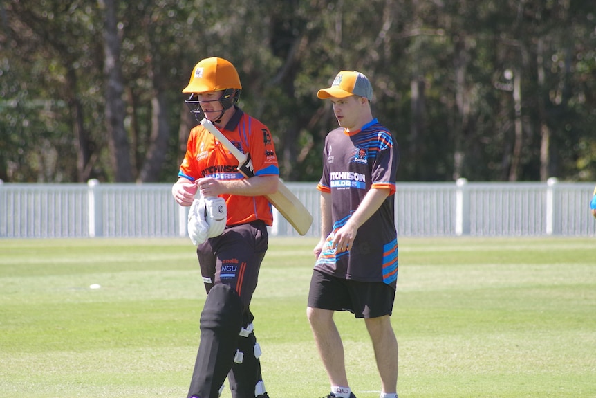 Two men walking. One wearing a cricket helmet and pads and holding a cricket bat and gloves. 