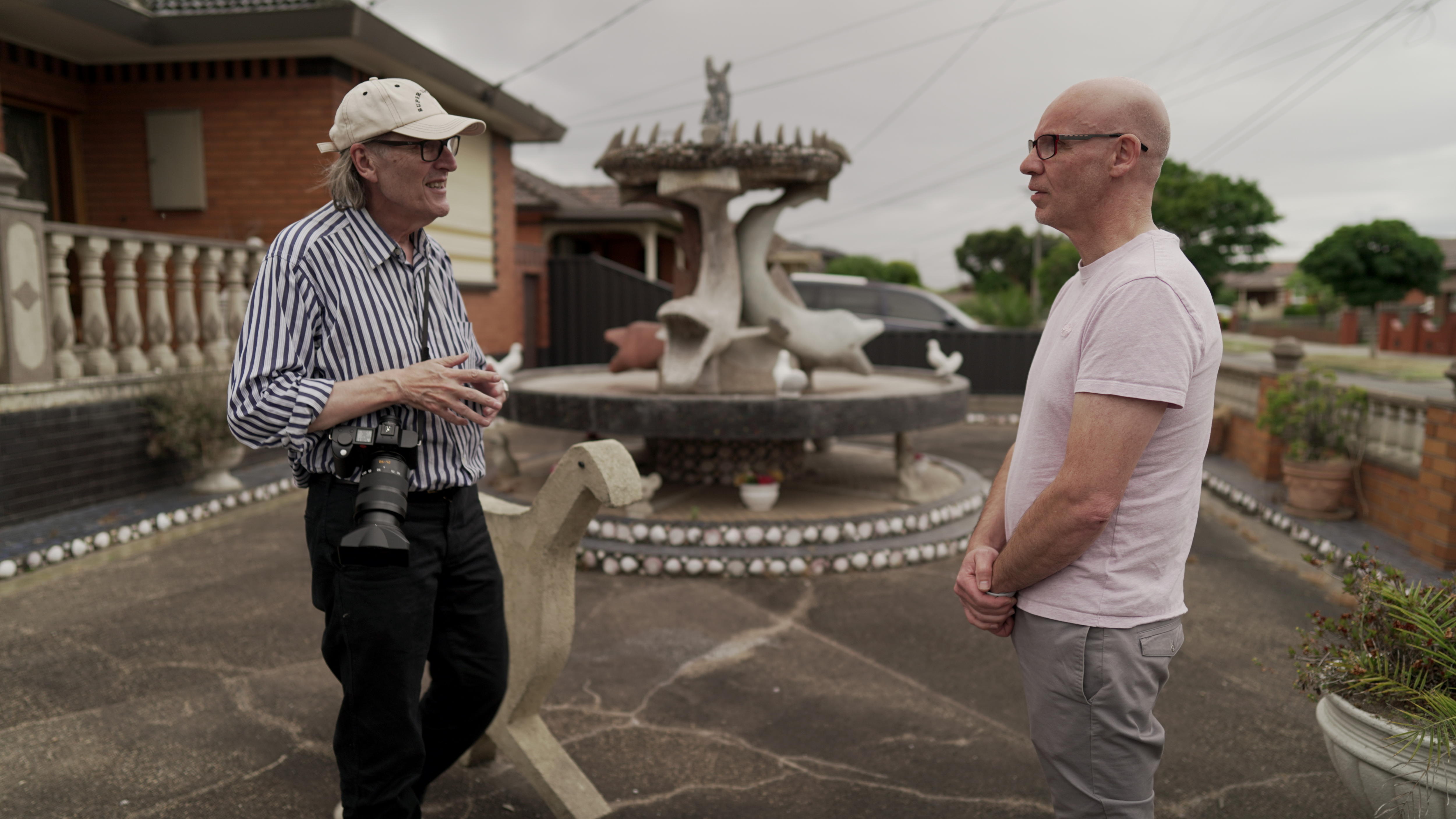 Two men are pictured talking in a front yard, behind them is a large fountain with concrete dolphins