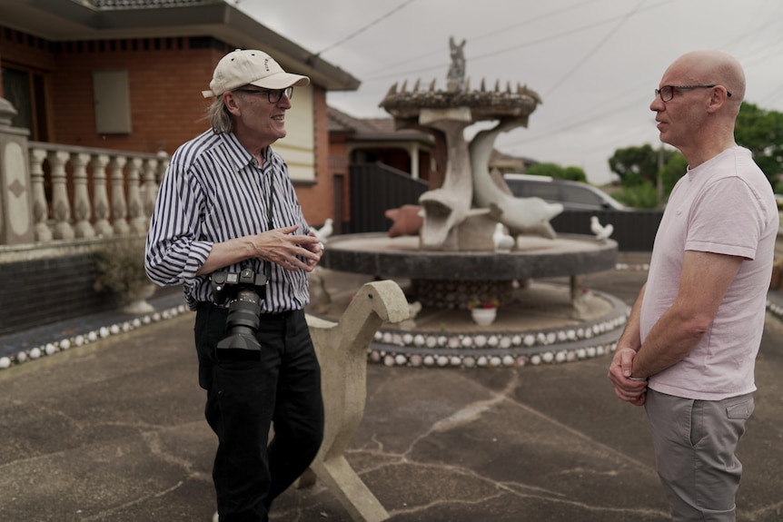 Two men are pictured talking in a front yard, behind them is a large fountain with concrete dolphins