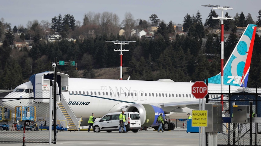 A white Boeing 737 jet is parked on the tarmac with workers and cars parked around its base with houses on hills in background.