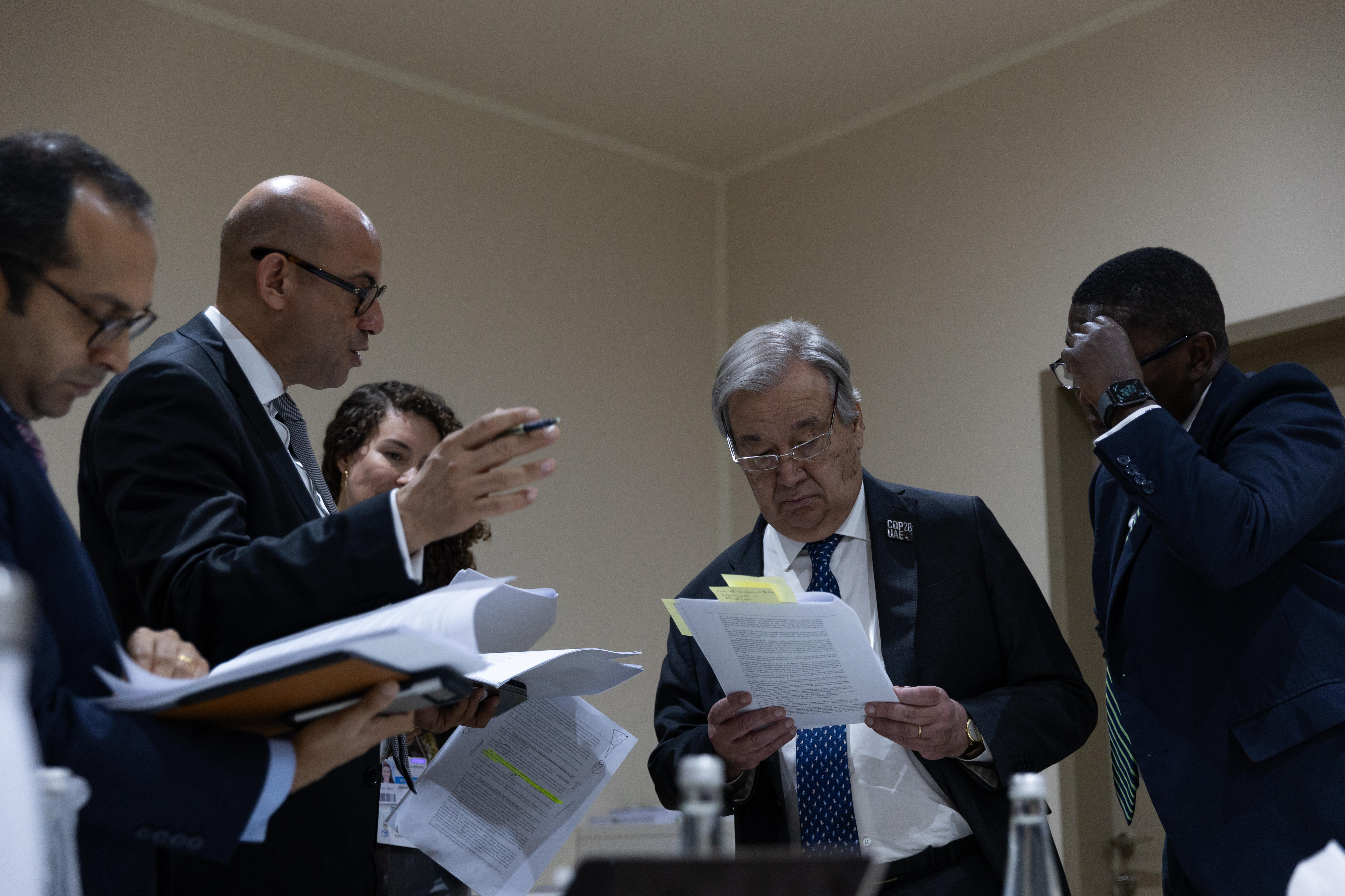 António Guterres stands reading draft text, annotated with sticky notes, surrounded by other people in suits discussing. 