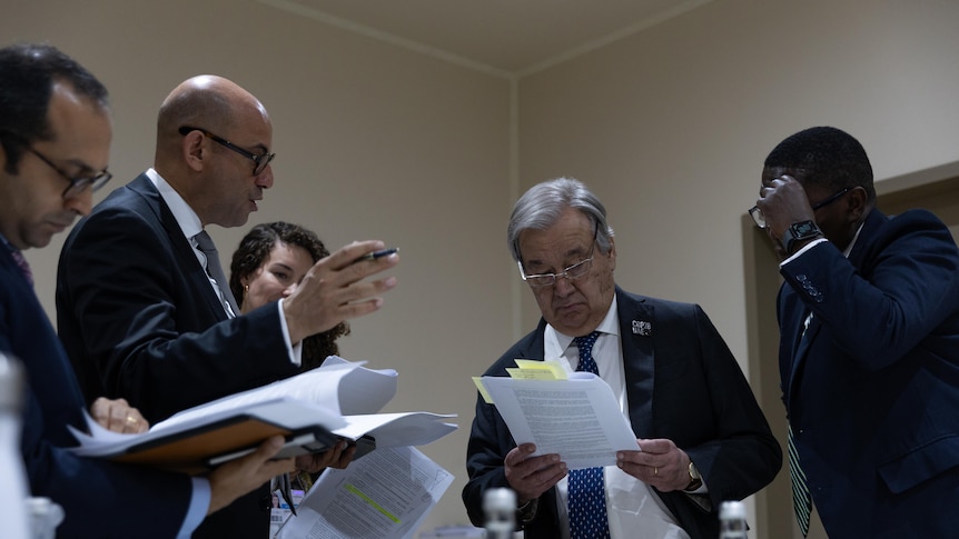 António Guterres stands reading draft text, annotated with sticky notes, surrounded by other people in suits discussing. 