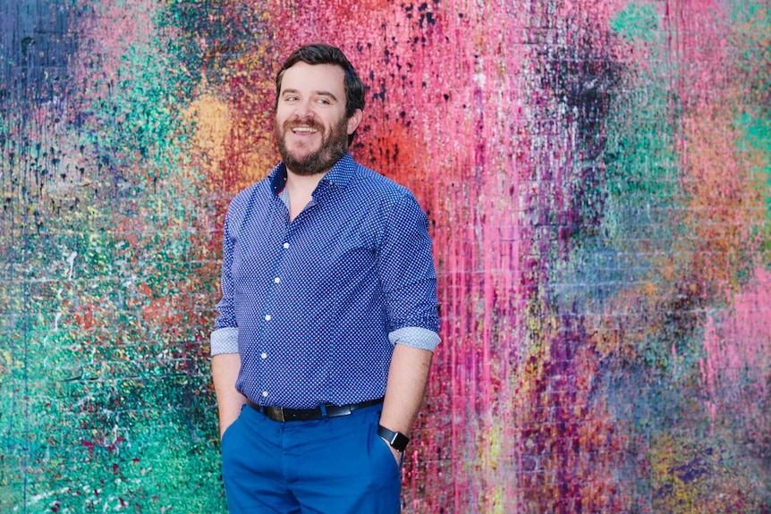 Williams, a man in his 40s, wearing all blue, smiles in front of a wall emblazoned with splattered, multi-coloured paint.