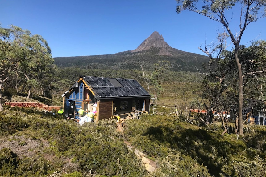 A modern hut under construction by a team of builders in a remote setting with Barn Bluff in the distance