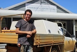 A man leaning against the back of a ute outside a general store.