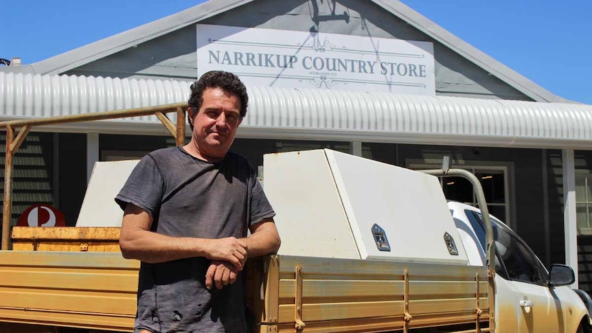 A man leaning against the back of a ute outside a general store.