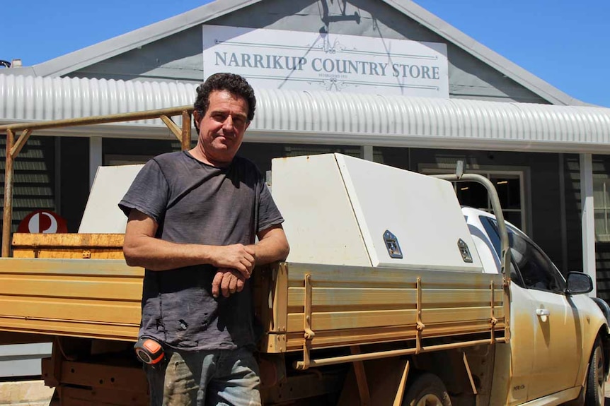 A man leaning against the back of a ute outside a general store.