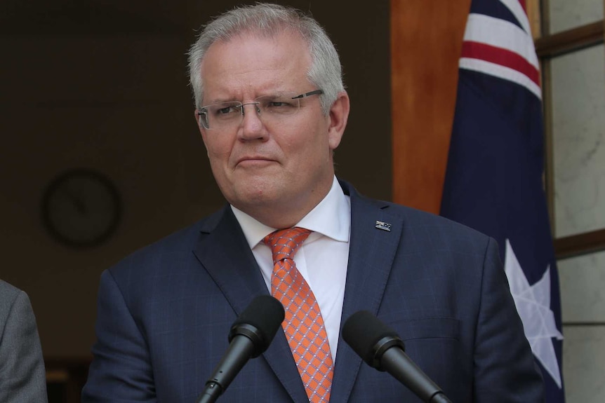 The Australian Prime Minister stands at a lectern in front of the national flag, wearing a blue suit and red tie.