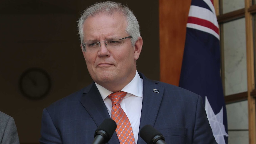 The Australian Prime Minister stands at a lectern in front of the national flag, wearing a blue suit and red tie.