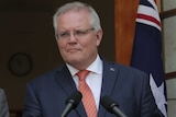 The Australian Prime Minister stands at a lectern in front of the national flag, wearing a blue suit and red tie.