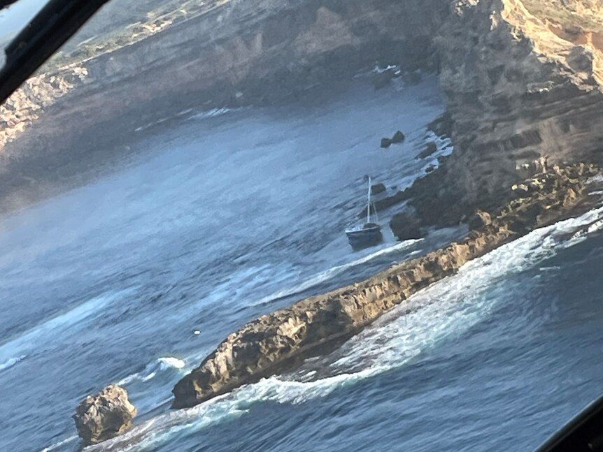 A yacht dangerously close rocks at the base of a massive cliff.