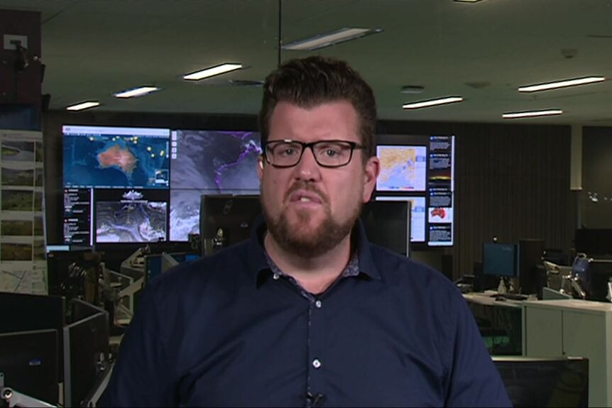 A man with a beard and glasses stands in a room with weather maps on digital screens in the background.