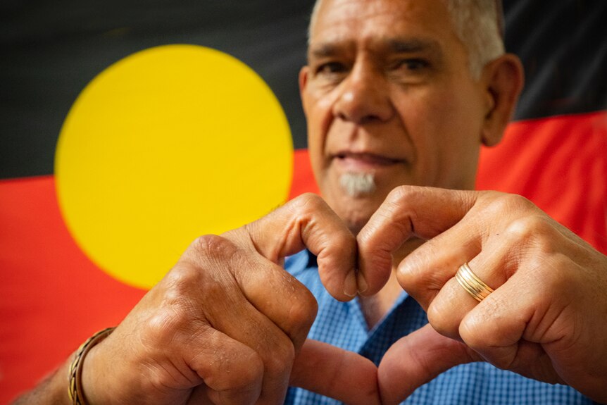 A man stands in front of the Australian Aboriginal flag, forming a heart with his hands.