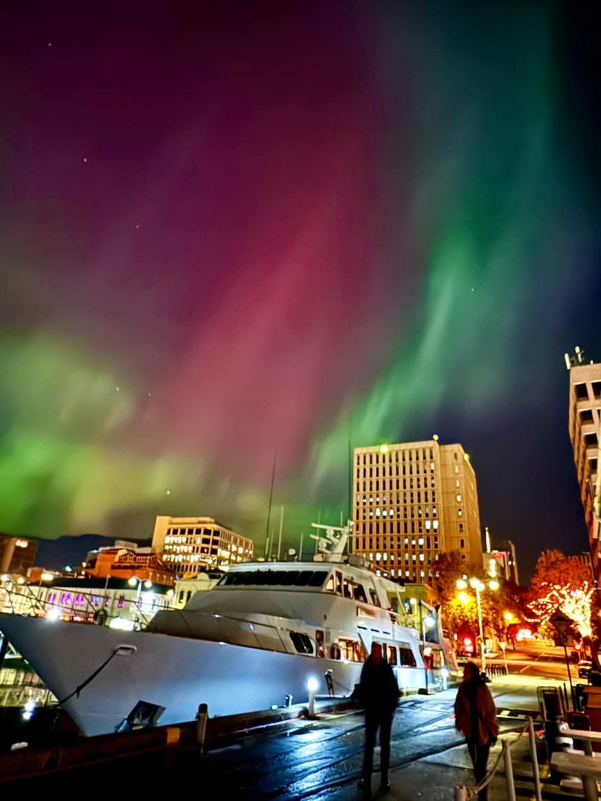 A green and pink aurora is seen behind a large boat docked in a city