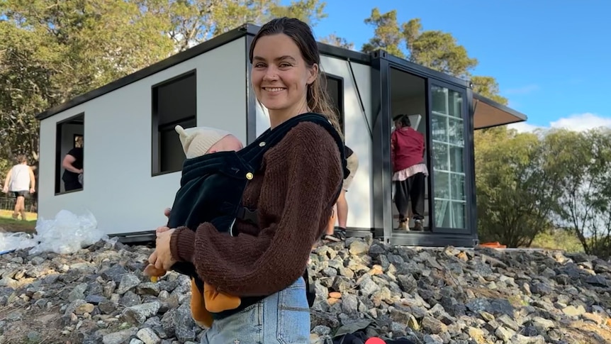 A woman stands in front of a small home on a hill holding her baby