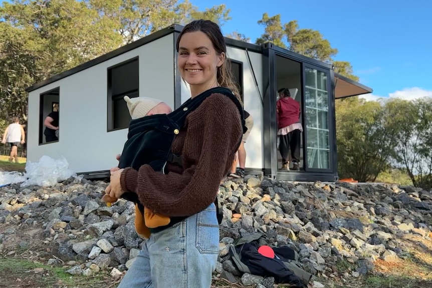 A woman stands in front of a small home on a hill holding her baby