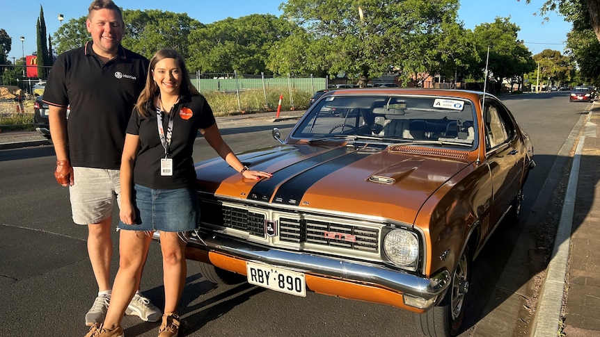 man and woman in black polo shirts stand in front of a brown 1969 Holden Monaro