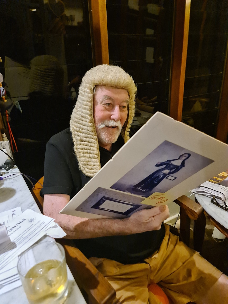 A elderly man, wearing a judge's wig, sitting on a chair and looking at a historical photo of a judge.