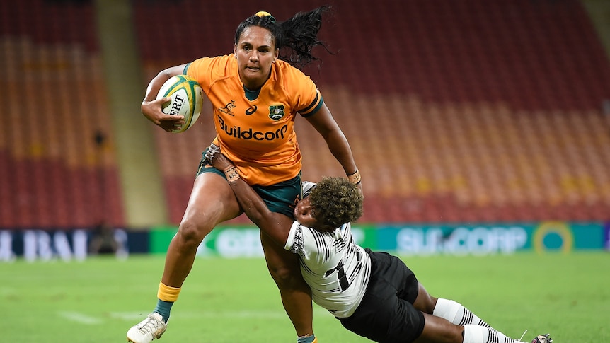 A Wallaroos player holds the ball with her right hand as she tries to fend off a Fijian opponent with her left.