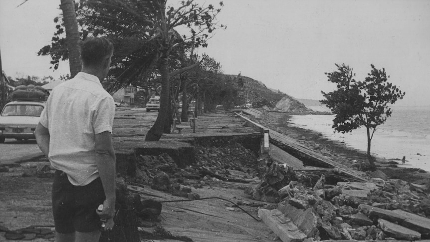 A black and white image of a damaged road and beach, with a man standing, holding an old-fashioned film camera.