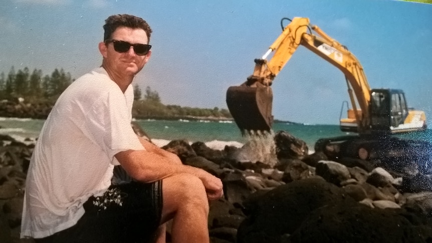 A large yellow excavator on dark grey, basalt rocks at the beach. A man appears small standing beside it.