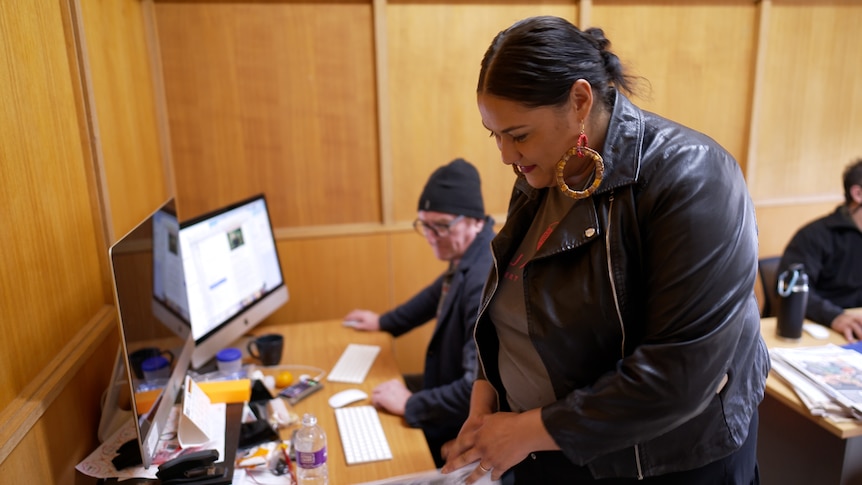 A woman in an office wearing a black leather jacket leaning over to read a newspaper