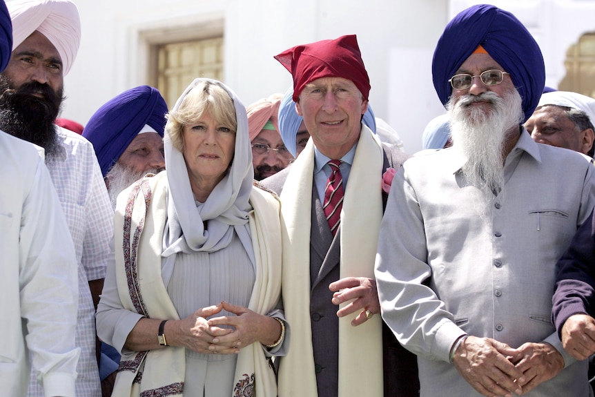 King Charles and Camilla wearing head coverings, next to Sikh man in turban, in India.