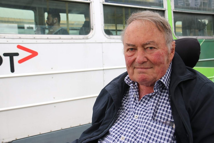 Tom Roper is dressed in a checked shirt and jacket as he sits in his wheelchair in front of a tram.