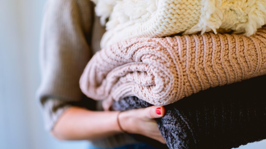 A woman holds a stack of three knitted blankets, close up shot of just her right arm holding the folded blankets