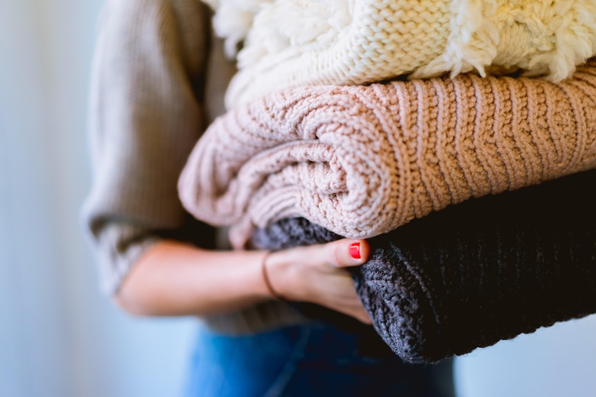A woman holds a stack of three knitted blankets, close up shot of just her right arm holding the folded blankets