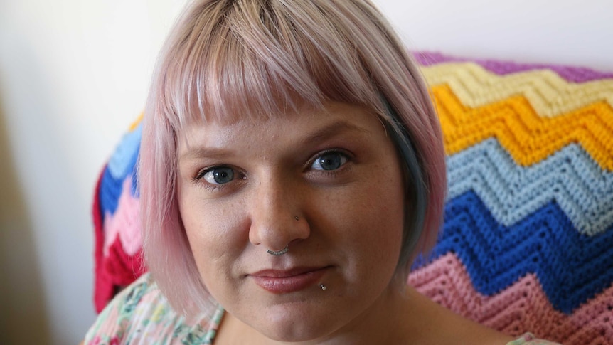 A young woman with pink-blonde hair and blue eyes look sits on a couch in her home.