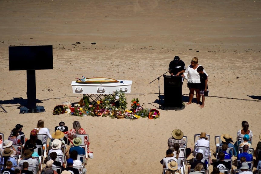 Family members speaking at a funeral service held on a beach.