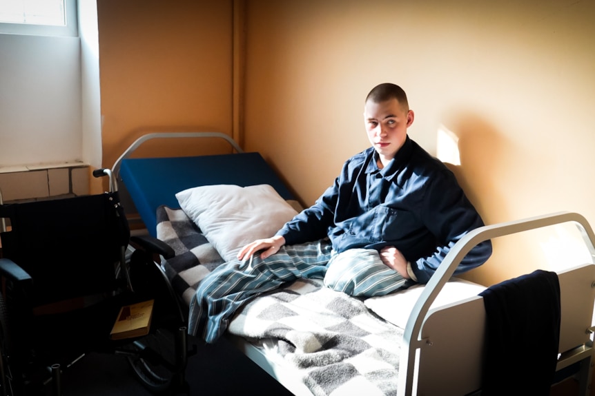 A young man, who has had both legs amputated, sits upright in a prison bed.