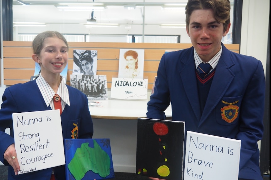 Abbey (left) and Hudson (right) in school uniform, holding up artwork in front of table with more artwork about Nida Lowe